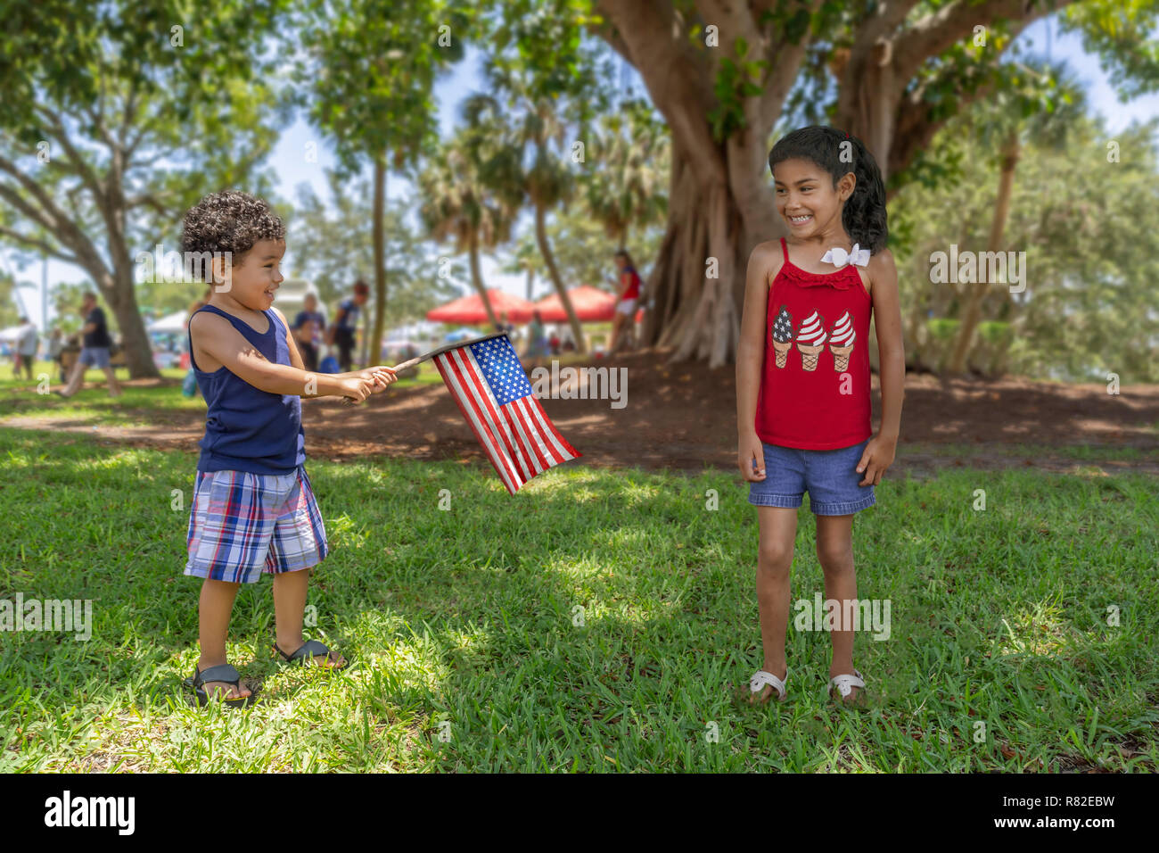 Ispanico fratello e sorella gioca alla comunità del parco sotto l'ombra di un grande albero come egli tiene la bandiera americana attaccato ad un bastone. Foto Stock