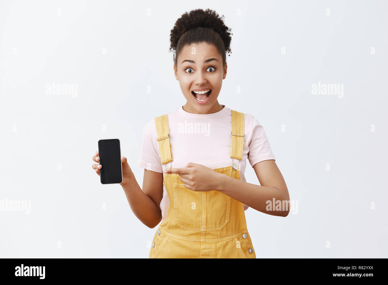 Prezzo incredibile per telefono incredibile. Stupiti ed eccitati African-American attraente ragazza con i capelli pettinati in tuta gialla, rivolta a sinistra a smartphone, che mostra lo schermo del dispositivo a telecamera Foto Stock