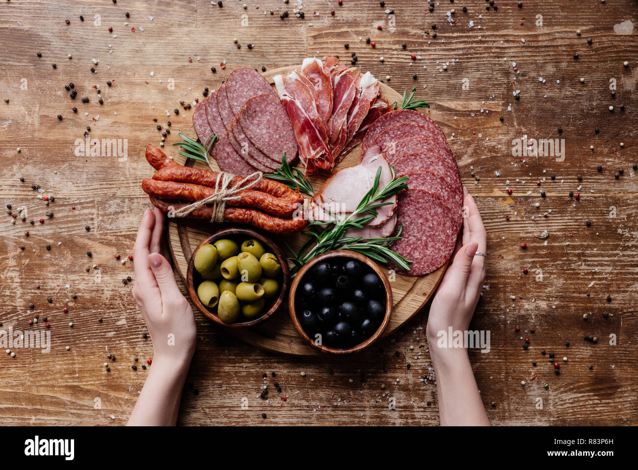 Vista dall'alto di mani femminili holding round tagliere con olive in due ciotole e fette di salame, prosciutto crudo e prosciutto su un tavolo di legno con sparse pe Foto Stock