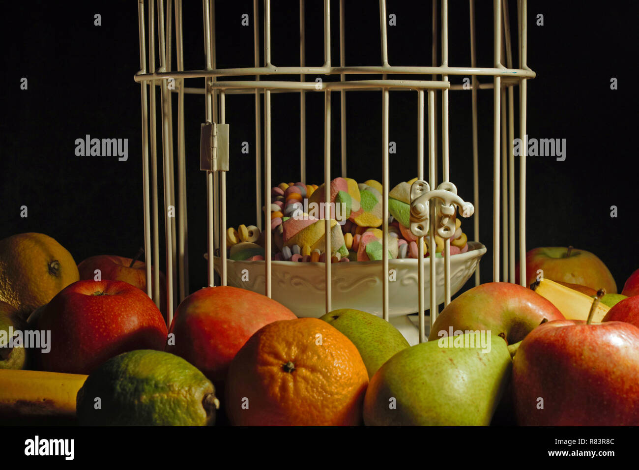 Mele, pere, mandarini e banane e limoni sono disposte intorno a un bianco di bird la gabbia contenente una ciotola di caramelle. Isolato su sfondo nero. Foto Stock