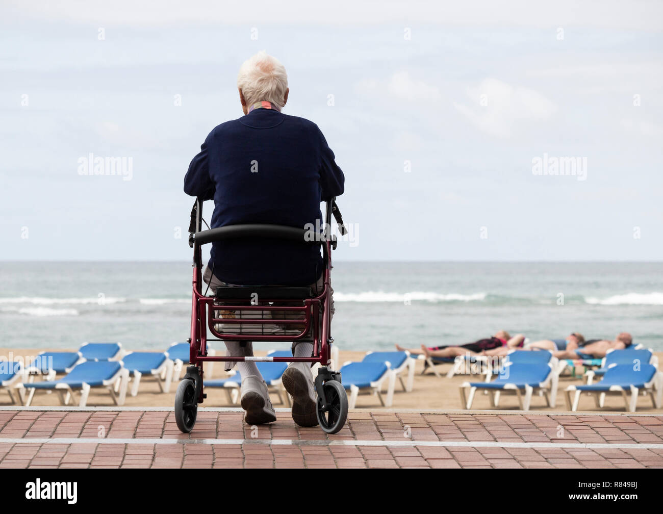 Anziani uomo seduto sulla ruota walker/rollator affacciato sulla spiaggia come le persone sole. Foto Stock