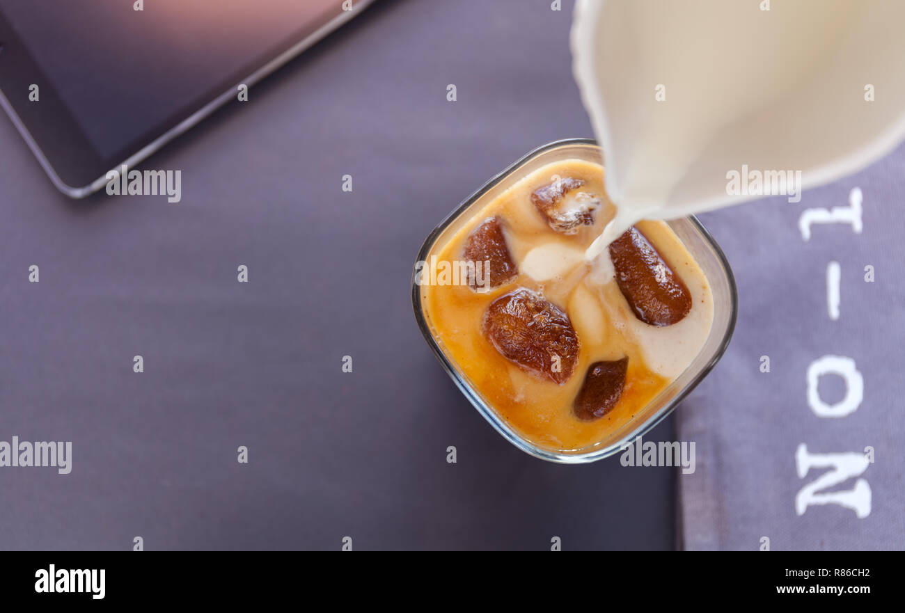 Versare il latte in un bicchiere di caffè ghiacciato i cubi su sfondo scuro. lettere numero 1 e tablet Foto Stock
