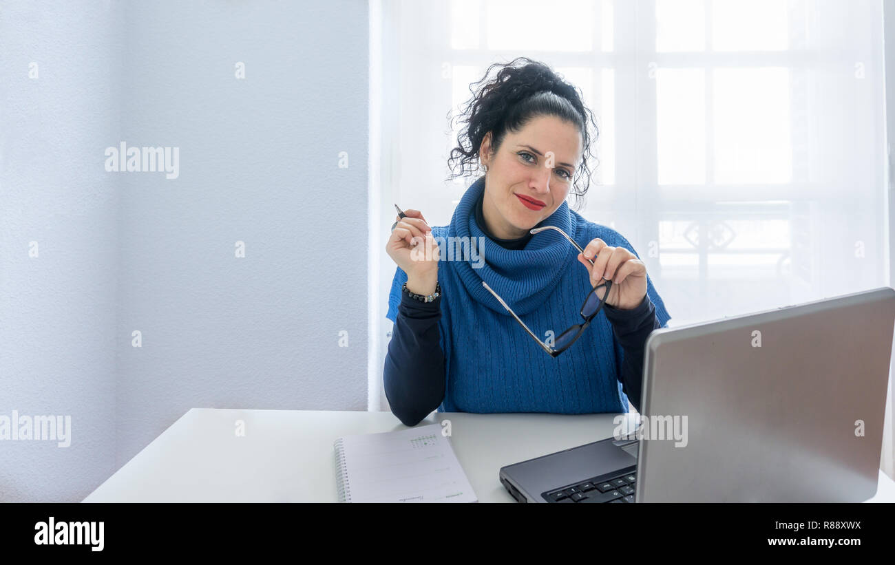 Ritratto di una donna normale con i capelli neri e la pelle bianca guardando la telecamera e tenendo gli occhiali dalla sua scrivania. In un ufficio con un ordine del giorno, laptop e notebook Foto Stock