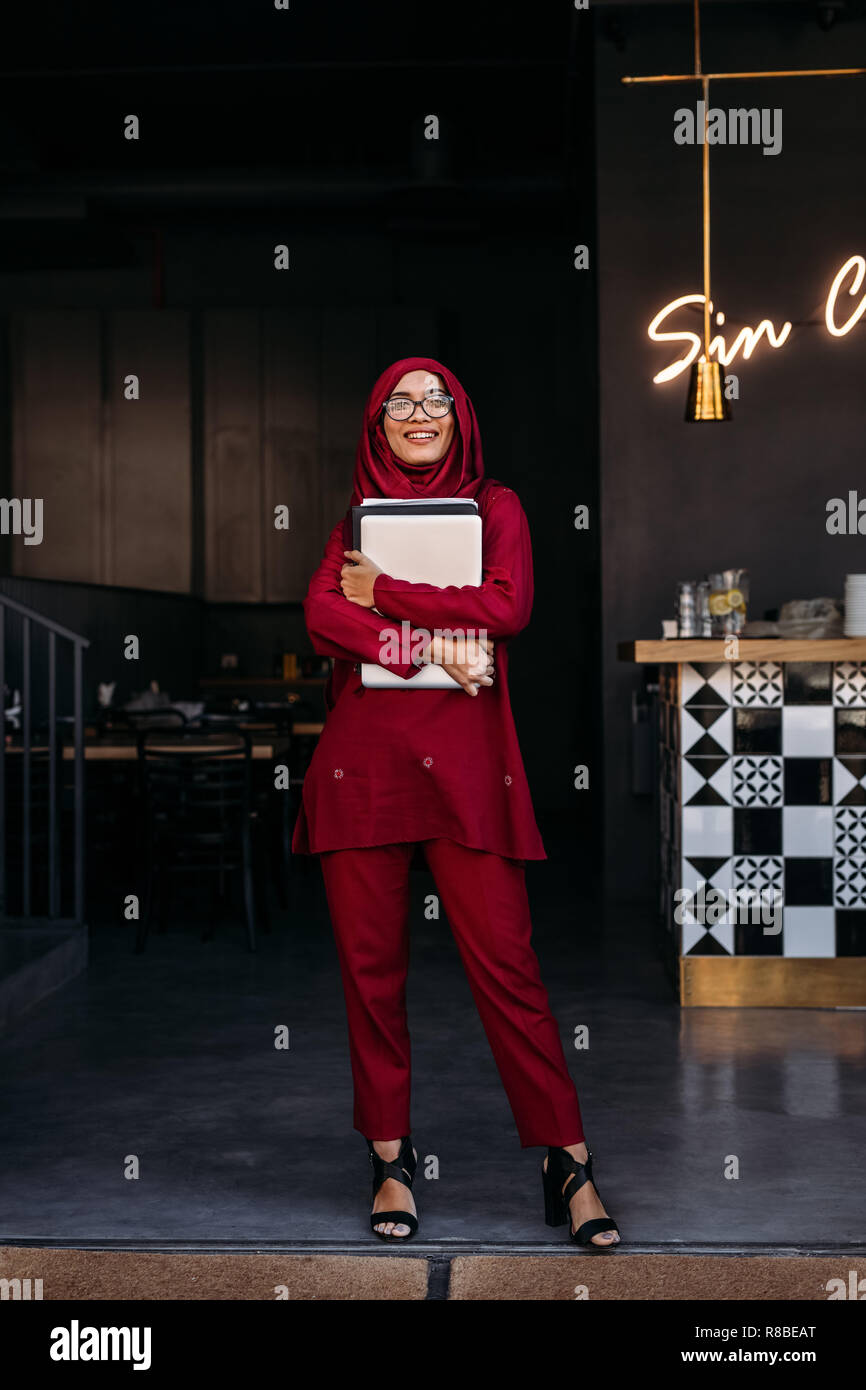 Per tutta la lunghezza della donna in hijab guardando la fotocamera mentre si sta in piedi in coffee shop azienda porta laptop. Business musulmano donna in piedi presso il ristorante porta. Foto Stock