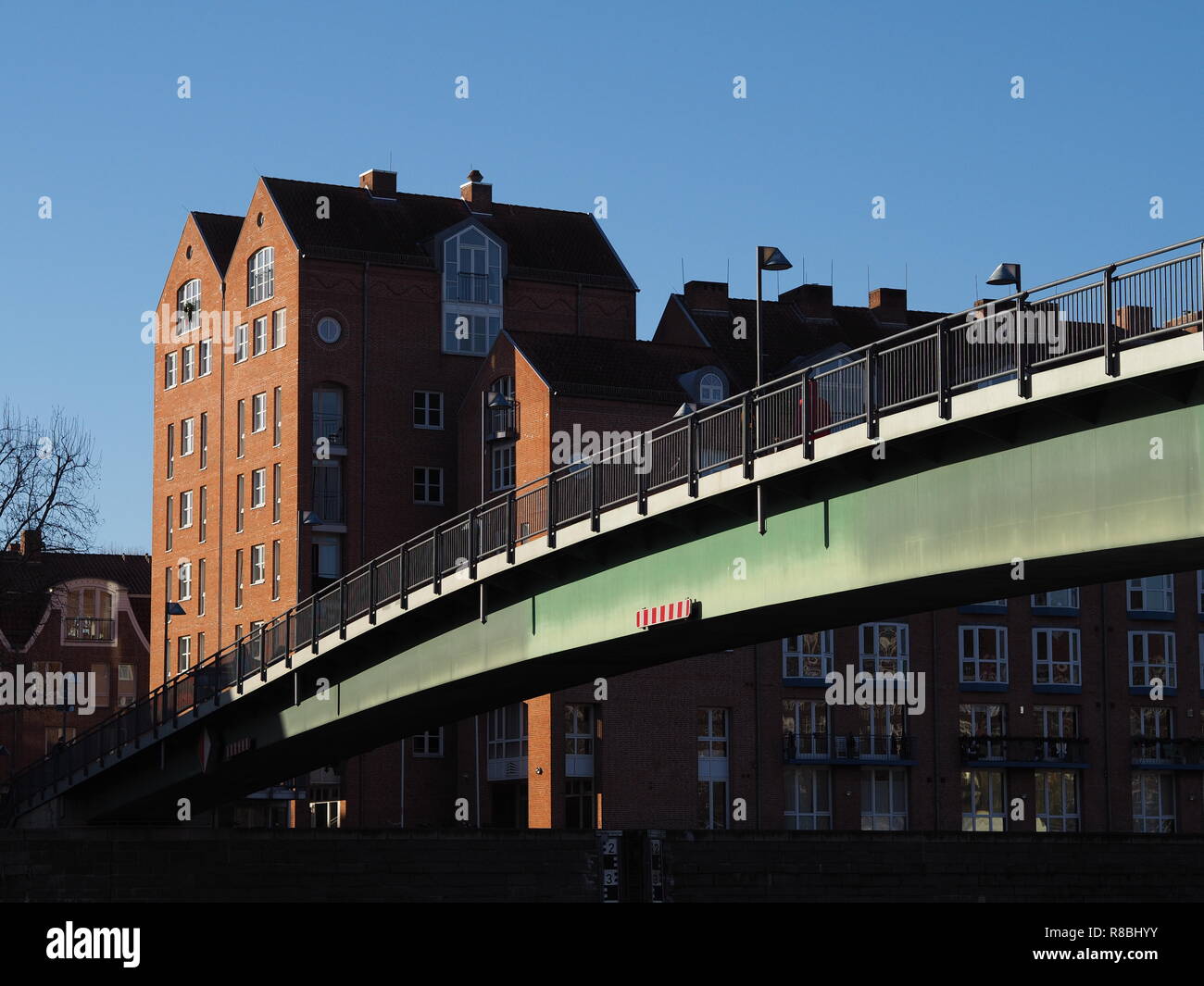 Bremen, Germania - acciaio ponte che attraversa il fiume Weser che conduce alla penisola Teerhof con mattoni rossi di edifici in background al di sotto di un blu limpido s Foto Stock