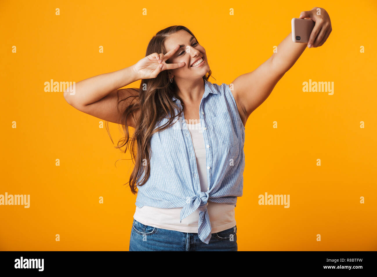 Allegro giovane donna in piedi isolato su sfondo giallo, prendendo un selfie con mano outsretched Foto Stock