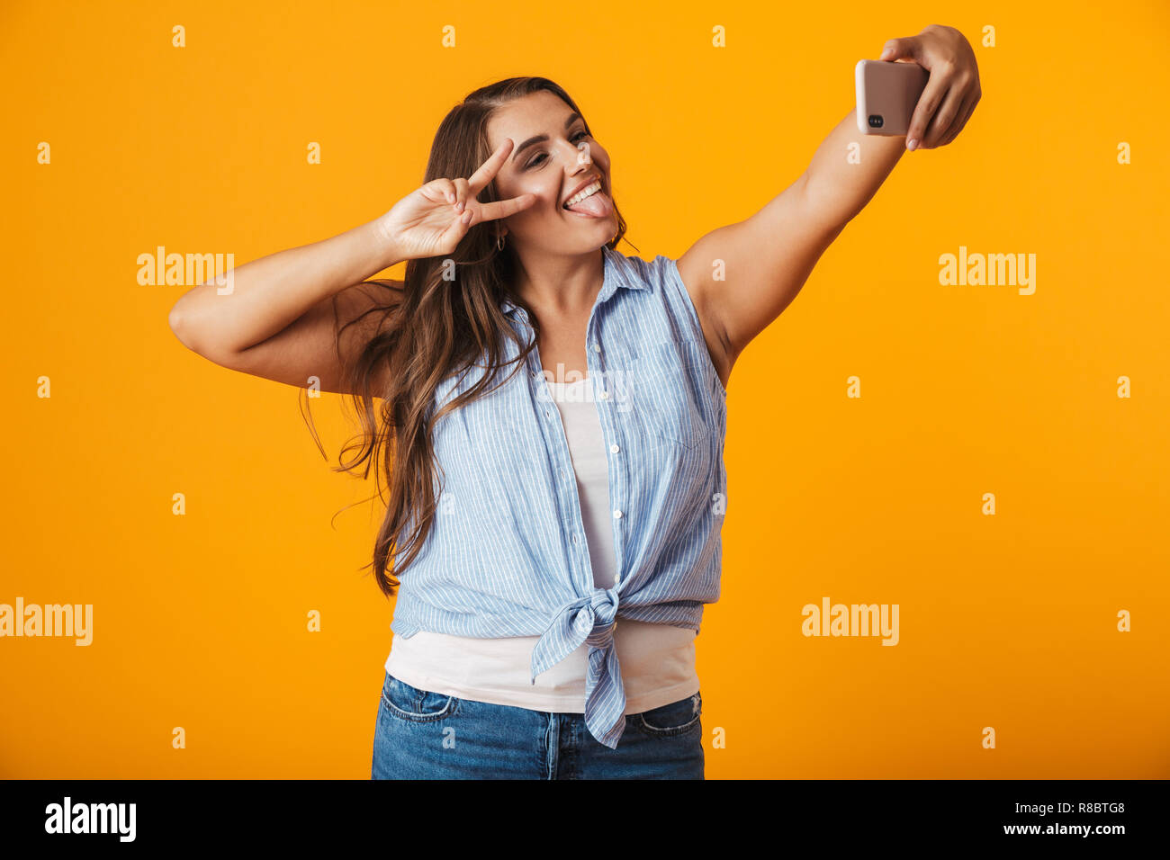 Allegro giovane donna in piedi isolato su sfondo giallo, prendendo un selfie con mano outsretched Foto Stock