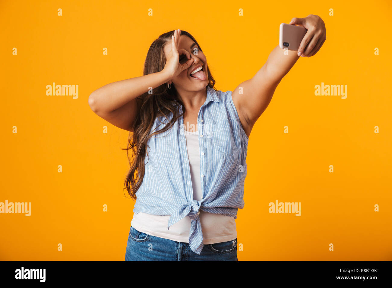 Allegro giovane donna in piedi isolato su sfondo giallo, prendendo un selfie con mano outsretched Foto Stock
