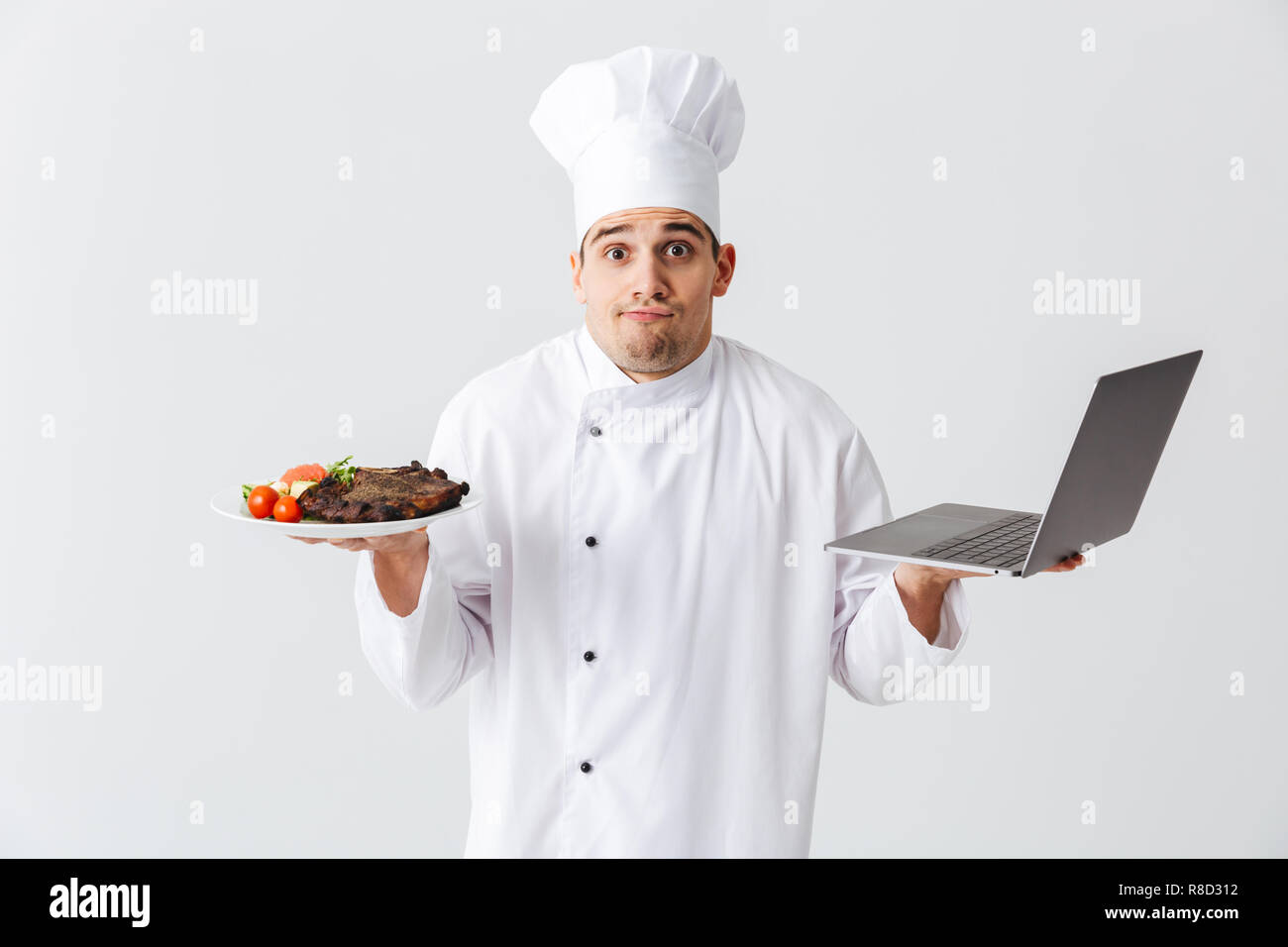 Confuso chef cuoco uniforme da indossare in piedi su sfondo bianco, tenendo il computer portatile, che mostra un piatto Foto Stock