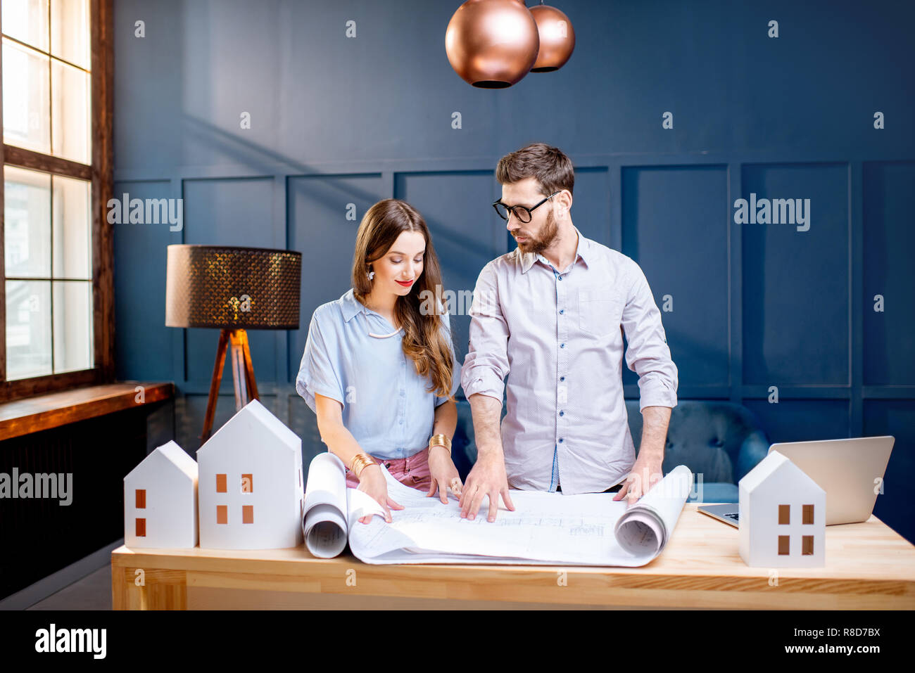 Giovane donna e uomo come gli architetti che lavorano con blueprint, laptop e modelli della casa in ufficio Foto Stock