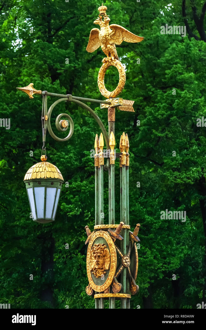 Lampione appeso nel giardino della città su un palo con una figura dorata di un aquila a due teste su uno sfondo di verdi alberi. Foto Stock
