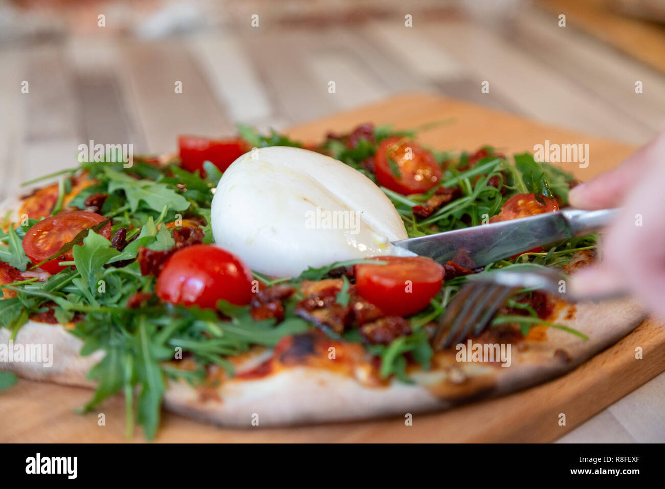 Mani tenendo una forchetta e un coltello per formaggio da taglio di una pizza su una tavola di legno con pomodorini, rucola e burrata formaggio su di un tavolo di legno e b Foto Stock