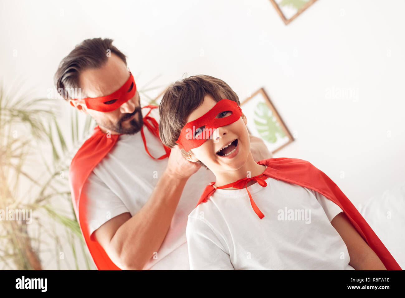 Padre e figlio in costumi superheroe a casa uomo permanente di nodo di chiusura sulla maschera del ragazzo che sorride allegro Foto Stock