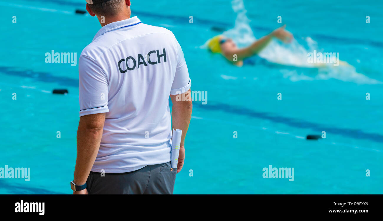 Vista posteriore di allenatori di nuoto, indossa una maglietta di pullman watiching la sua femmina nuotatore facendo dorso a livello locale piscina esterna Foto Stock