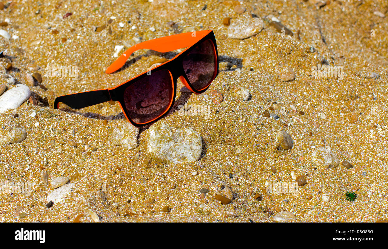 Gli occhiali da sole sono persi e giacciono sulla spiaggia in riva al mare. Nero con bordo arancione bicchieri sullo sfondo di giallo sabbia sulla riva. Gli occhiali da sole sono ri Foto Stock