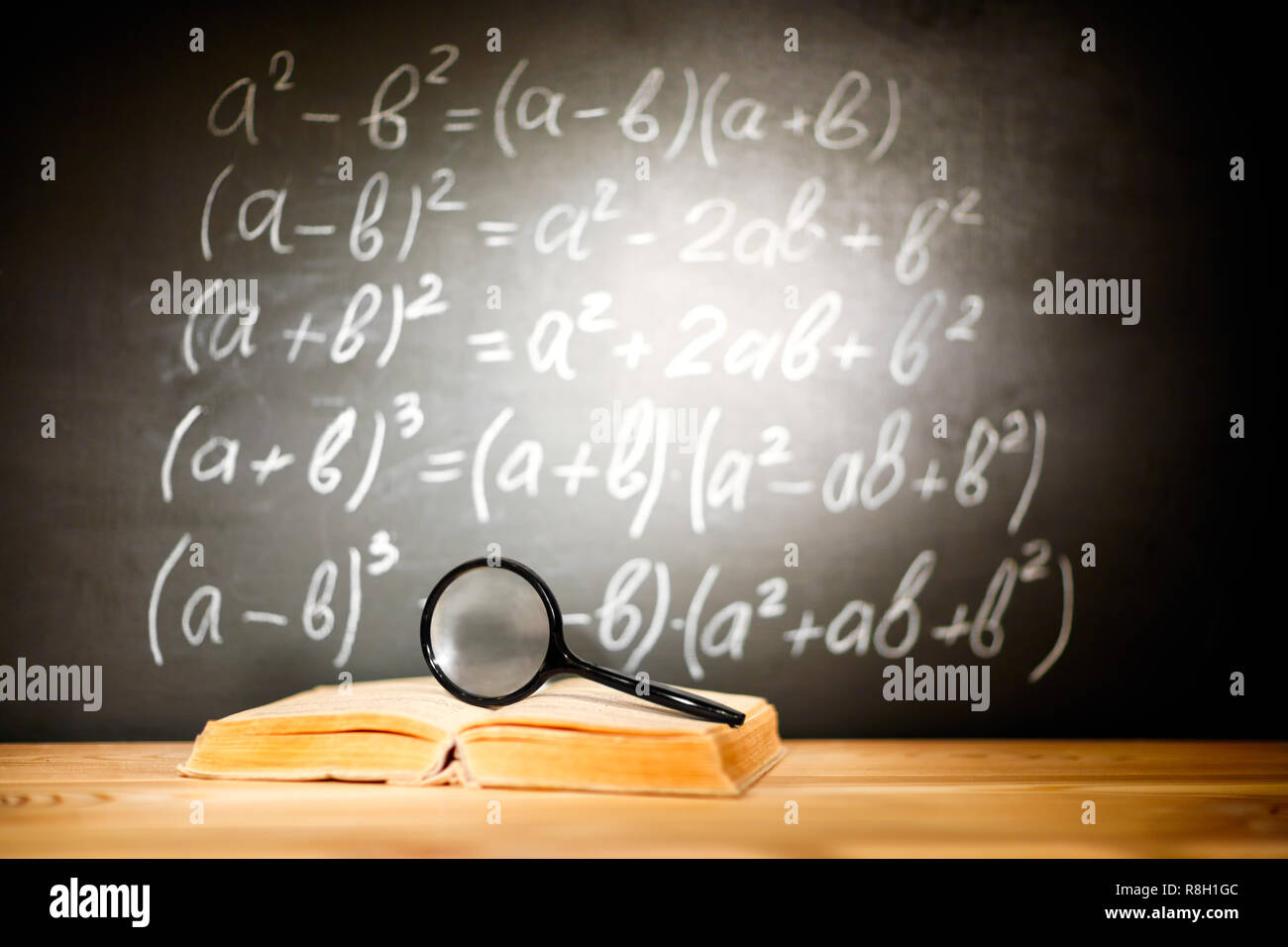 Vecchi libri scolastici e magnifier giacente su una tavola di legno a banchi di scuola di fronte a una lavagna nera con formule matematiche scuola. Il concetto di istruzione - il Foto Stock