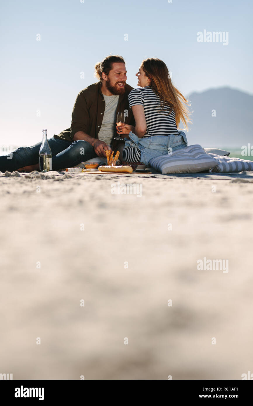 Bella giovane godendo di buon umore e giorno picnic sulla spiaggia. Basso angolo di visione di un uomo e di una donna seduta sulla spiaggia con cibo e bevande. Foto Stock