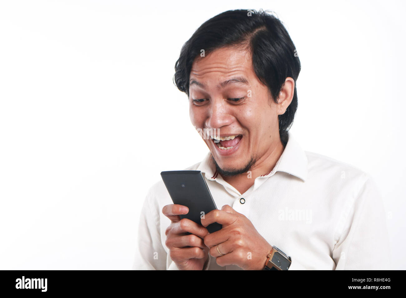 Foto immagine ritratto di un divertente giovane uomo asiatico sorpresi e sorridenti mentre cerca il suo smart phone. Tenendo il telefono con entrambe le mani durante la lettura della Messa Foto Stock