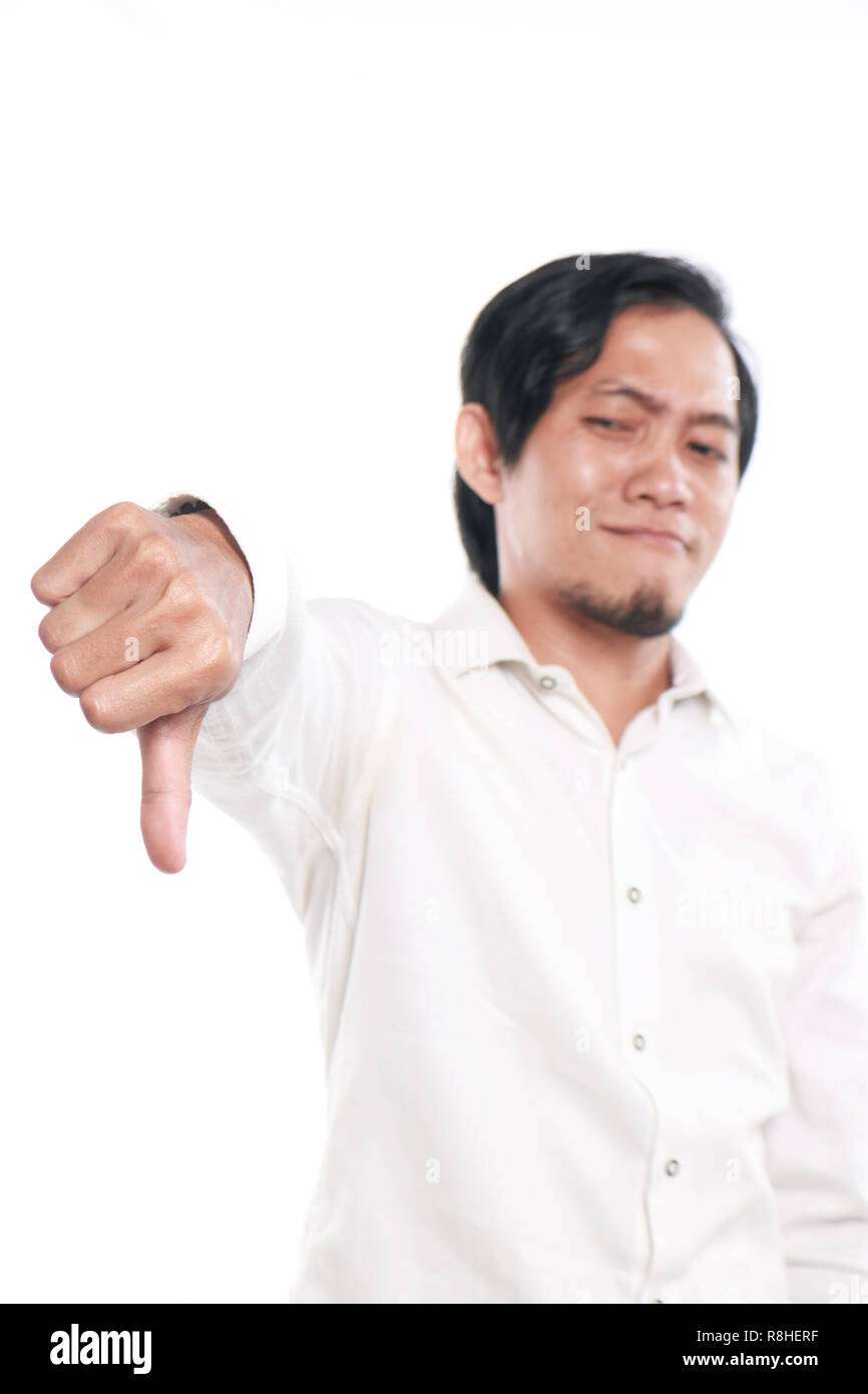 Foto immagine ritratto di un simpatico divertente giovane uomo asiatico che mostra il pollice verso il basso con gesto faccia beffarda, close up ritratto su sfondo bianco Foto Stock