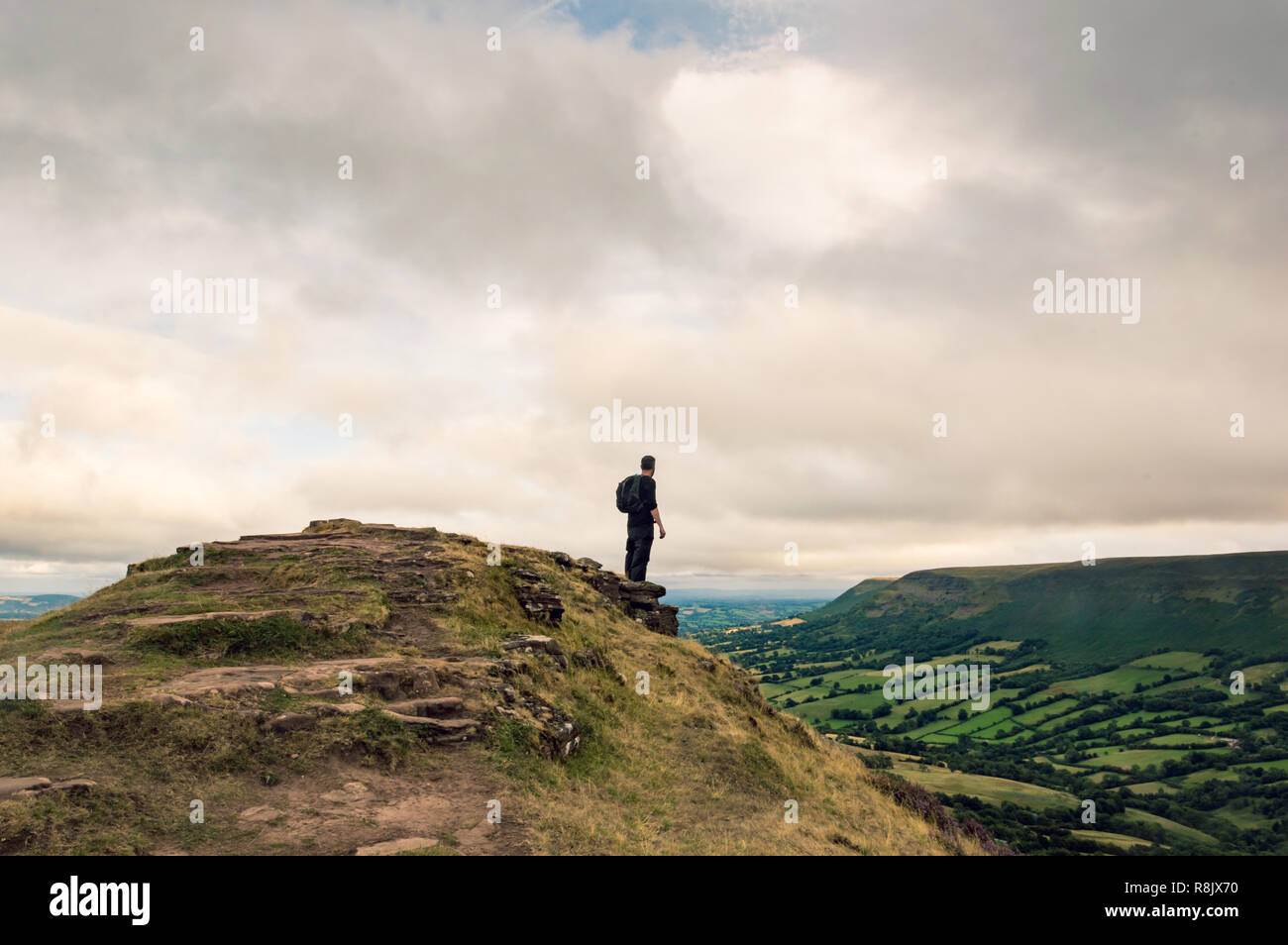 Un uomo con uno zaino guardando attraverso il paesaggio di campi e colline dalla sommità di una collina in inglese, confine gallese. Le montagne nere, il Galles. Foto Stock