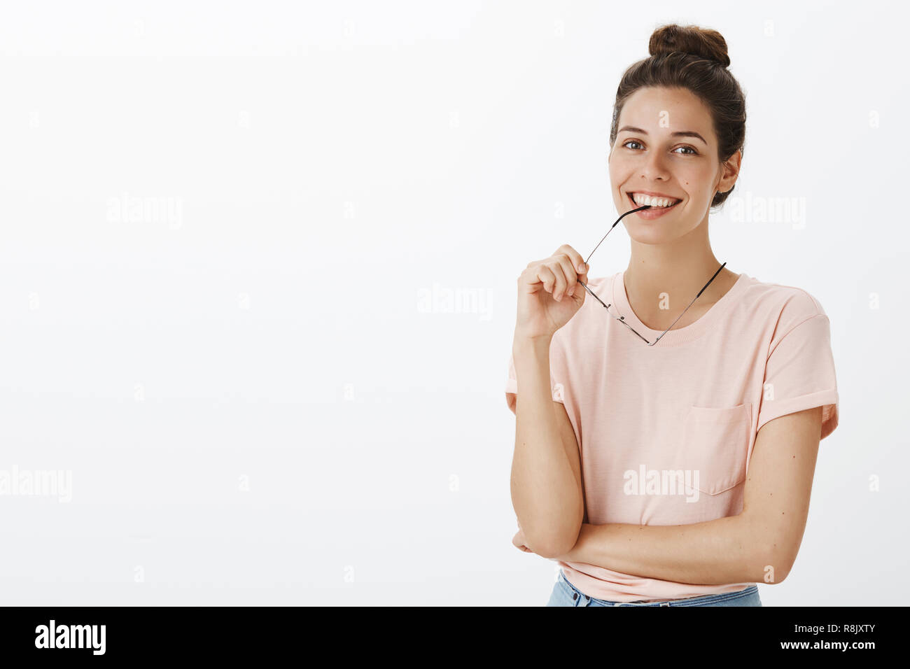 Vita-up shot di fiduciosa felice e abile team femminile stati mordere il telaio di occhiali come comportanti gioiosamente con auto-assicurato grin su sfondo grigio attraversando un braccio contro il petto Foto Stock