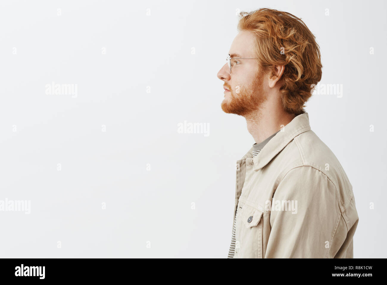 Colpo di profilo di calma bello e creative redhead modello maschile con capelli mossi e barba in vetri trasparenti e cappotto beige guardando a sinistra fiducioso in piedi in casual carefree pongono Foto Stock