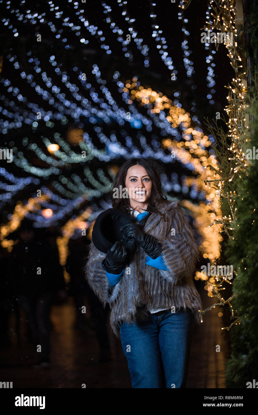 Piuttosto scuro ragazza dai capelli indossando una pelliccia, jeans blu, blu in alto e un cappello nero, sorridente, posa con i fiocchi di neve le luci di Natale all'aperto durante la notte ti Foto Stock