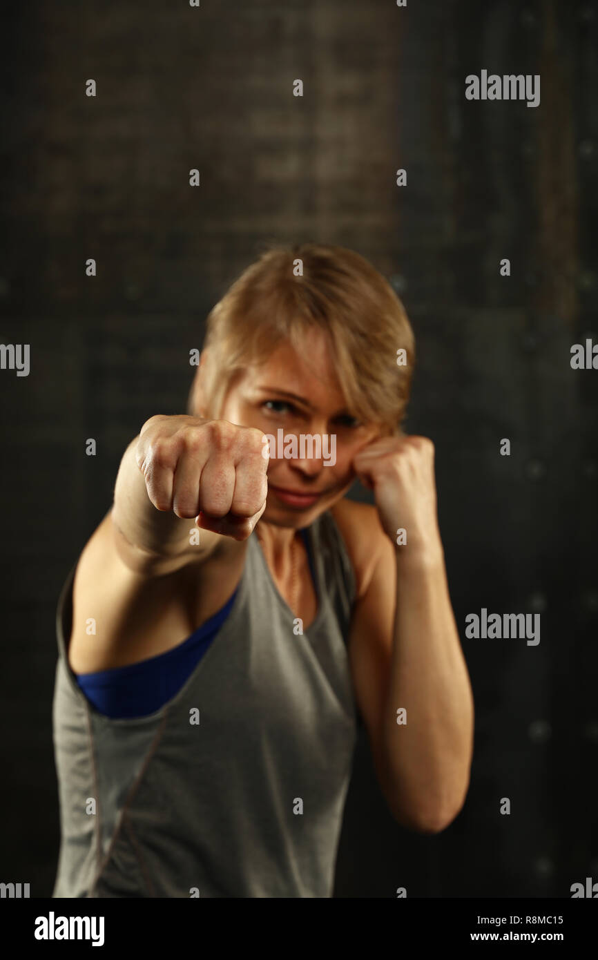 Close up front ritratto di una giovane età media athletic donna in abbigliamento sportivo in palestra su sfondo scuro, in piedi in atteggiamento di inscatolamento con mani e fis Foto Stock