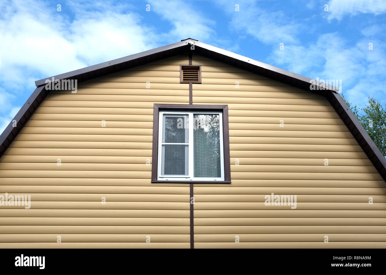 Piano superiore con finestra in plastica in country house parete ricoperta con schierandosi giallo e marrone e tetto metallico sul cielo blu con nuvole sulla giornata soleggiata vista frontale Foto Stock