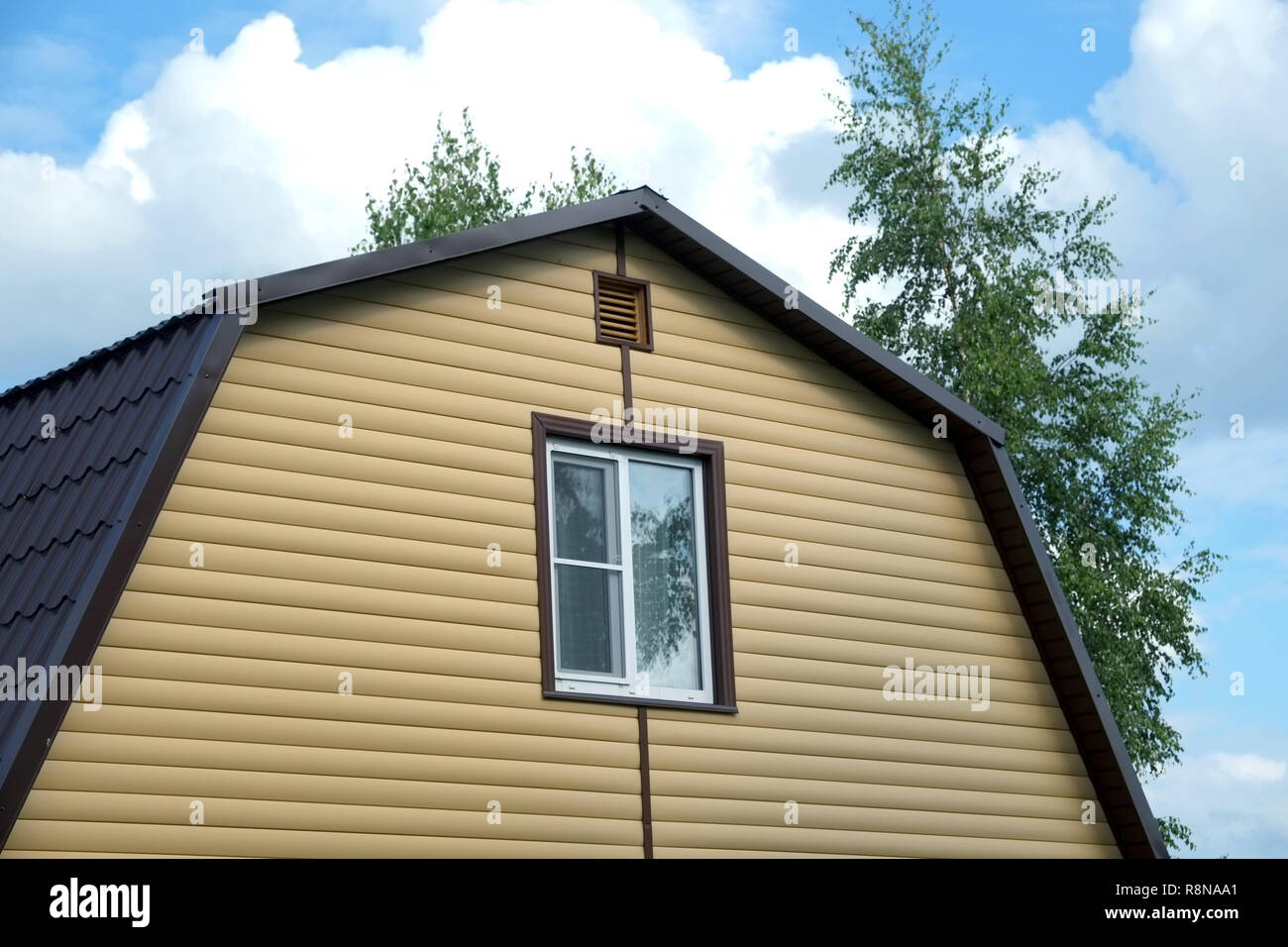 Piano superiore con finestra in plastica in country house parete ricoperta con schierandosi giallo e marrone e tetto metallico sul cielo blu con nuvole sulla giornata di sole Foto Stock