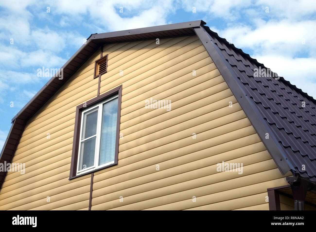 Piano superiore con finestra in plastica in country house parete ricoperta con schierandosi giallo e marrone e tetto metallico sul cielo blu con nuvole sulla giornata soleggiata vista laterale Foto Stock