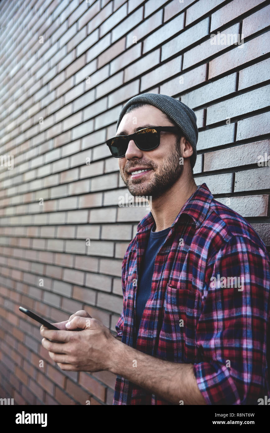 Hipster i messaggi sms app Telefono in strada di città su un muro di mattoni dello sfondo. Incredibile uomo tenendo lo smartphone nel smart casual in piedi. Urbano profe giovani Foto Stock