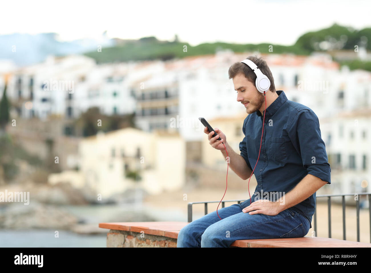 Uomo serio ascolto della musica da un telefono intelligente seduto su una battuta in una città costiera in vacanza Foto Stock