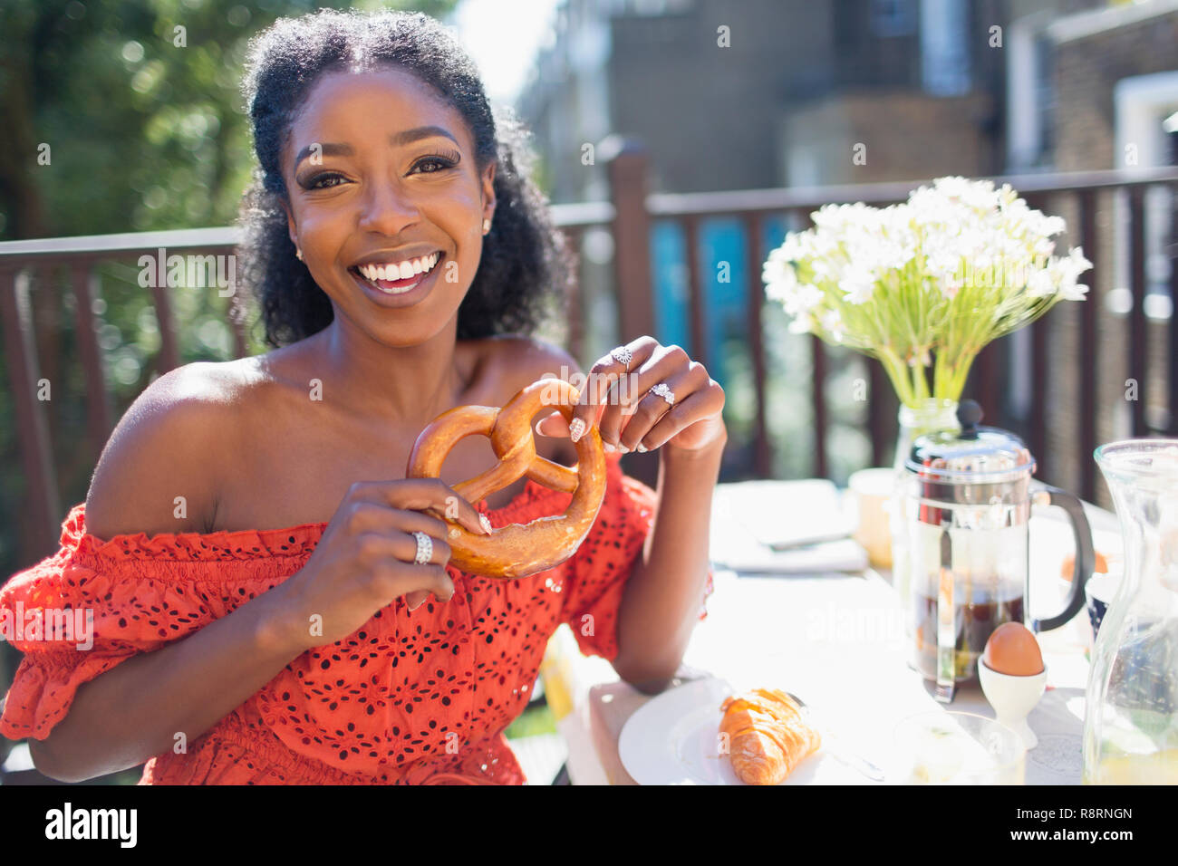 Ritratto sorridente, fiducioso giovane donna con pretzel gustando la prima colazione sul balcone soleggiato Foto Stock