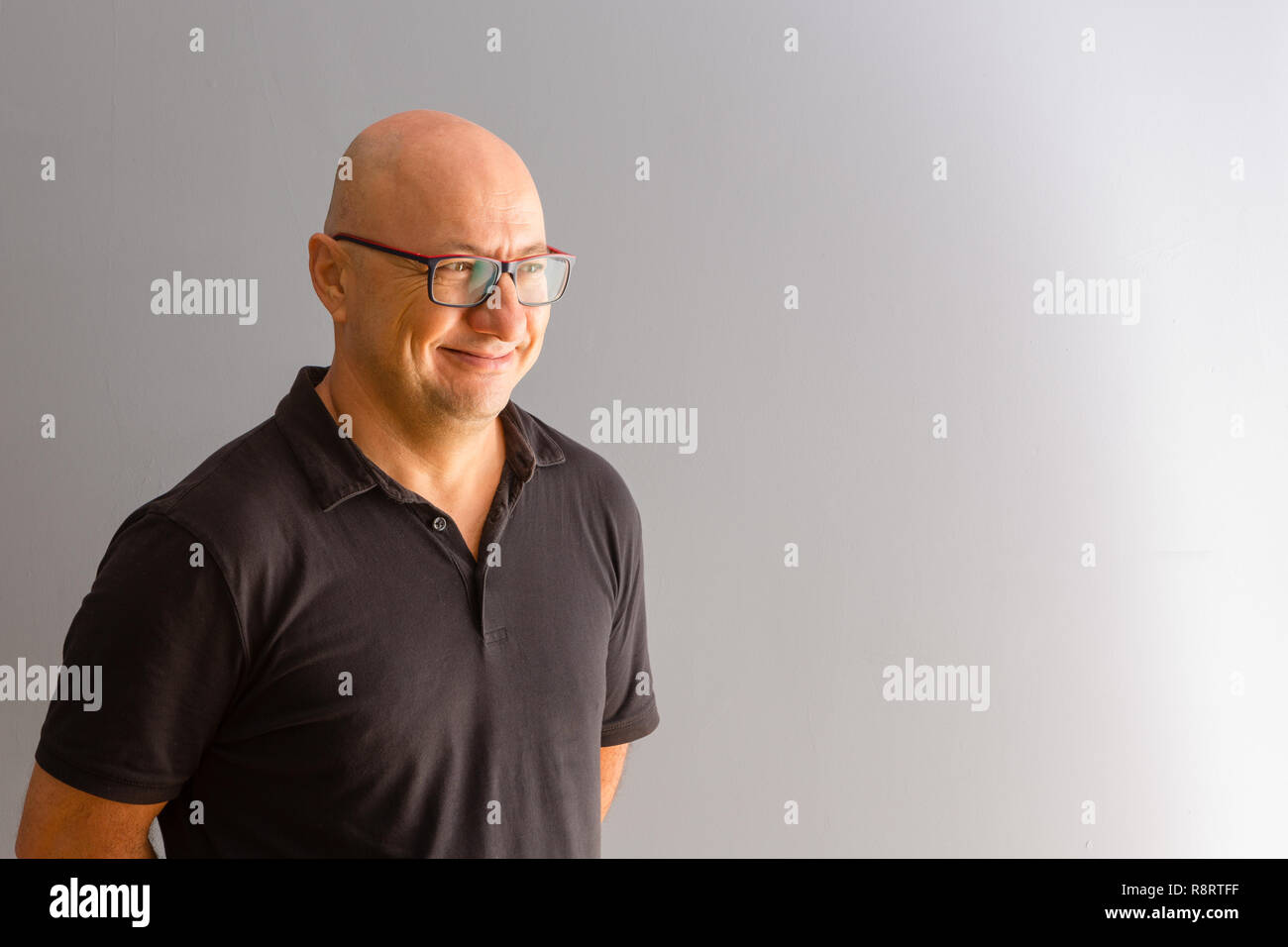 Adulto Uomo in grassetto in bicchieri e polo nero mezzo giro a mezza lunghezza ritratto, guardando al lato di grigio spazio copia e sorridente Foto Stock