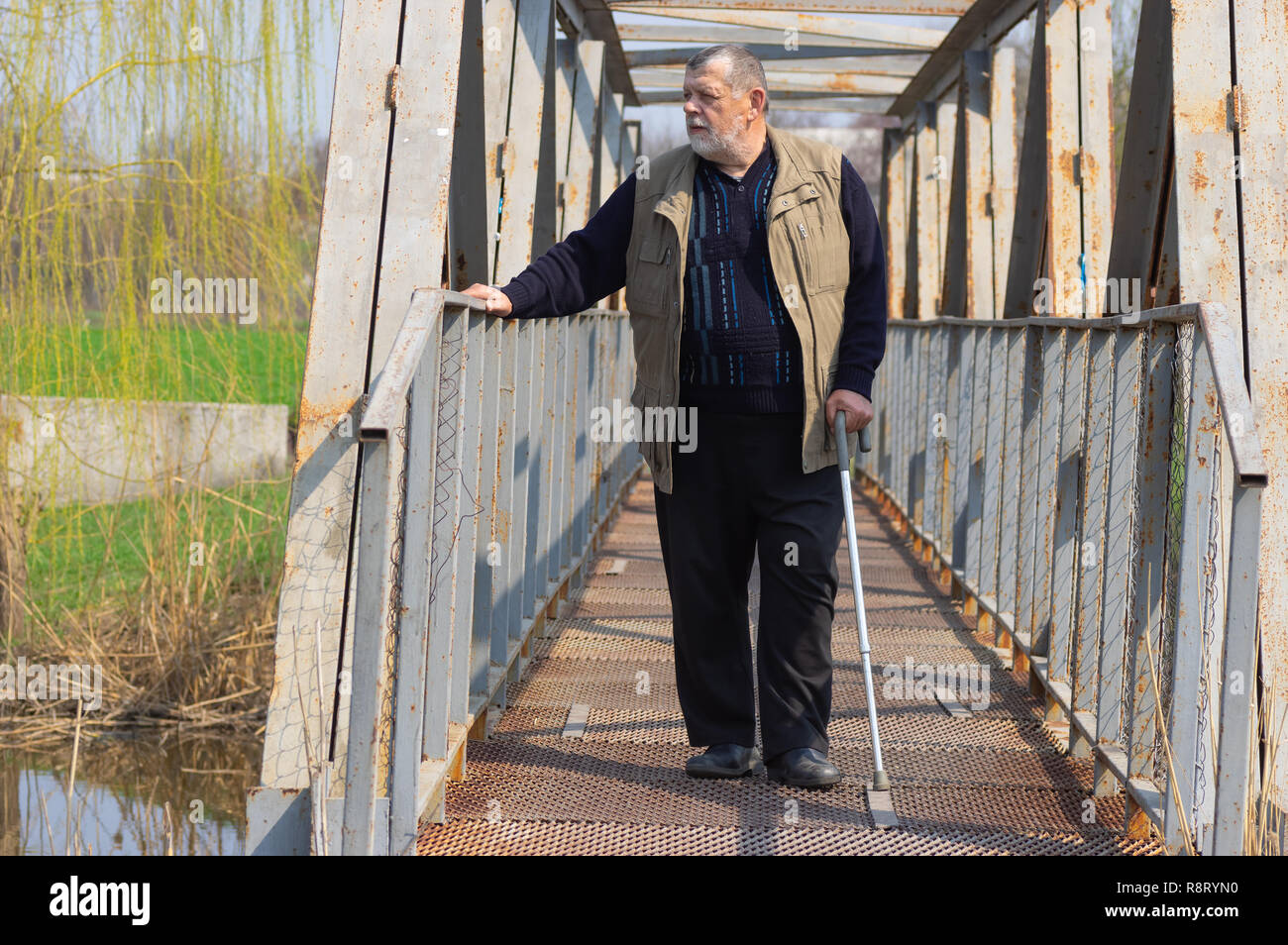Senior uomo con bastone in piedi su un ponte metallico oltre il piccolo fiume Foto Stock