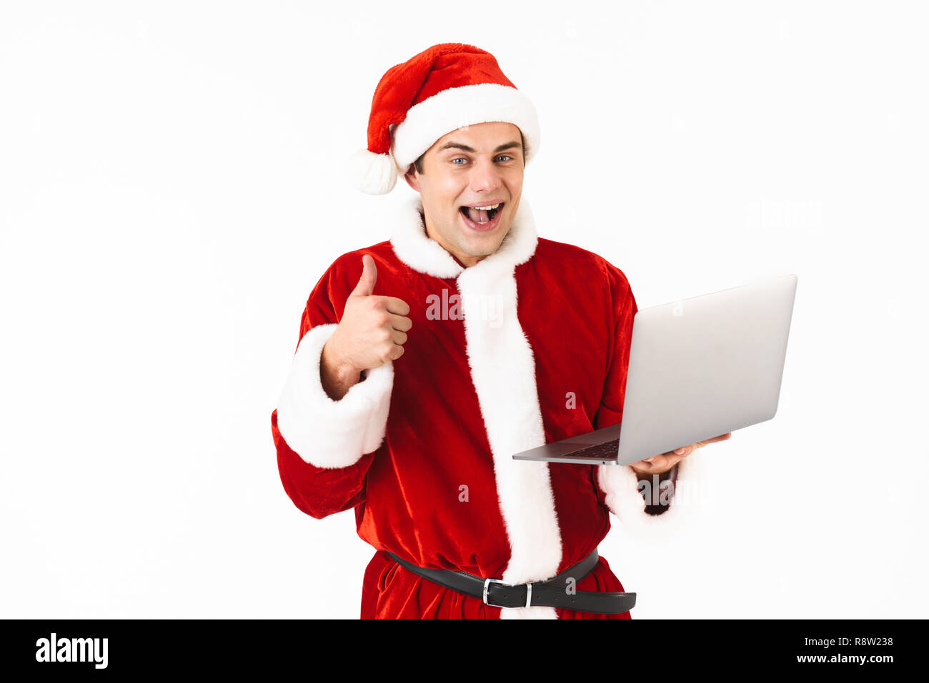 Ritratto di un allegro giovane uomo vestito di santa claus costume isolato permanente su sfondo bianco, tenendo il computer portatile, mostrando pollice in alto Foto Stock