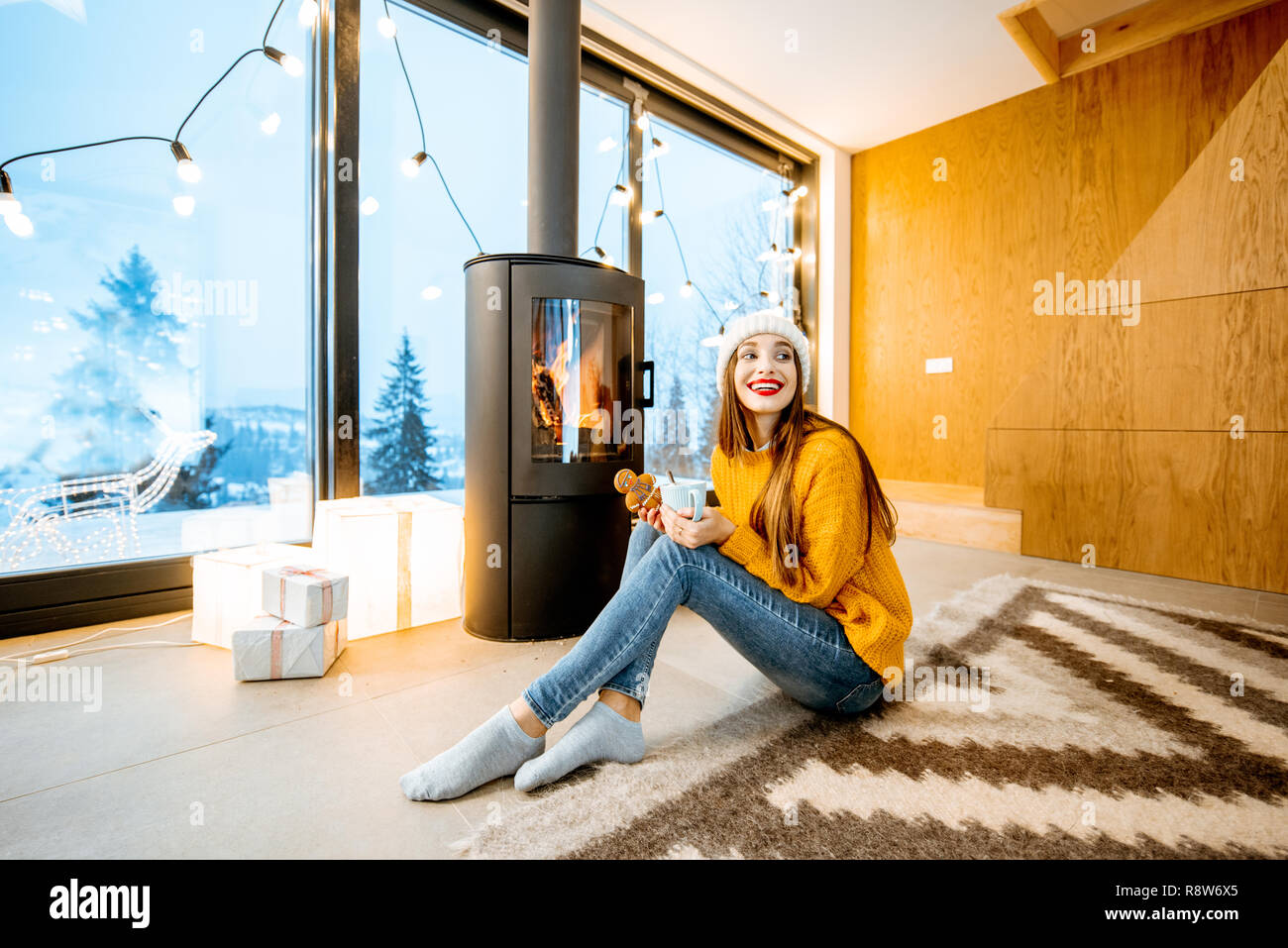 Ritratto di una giovane donna in un maglione e hat seduta con bevanda calda vicino al camino nella casa moderna in montagna Foto Stock