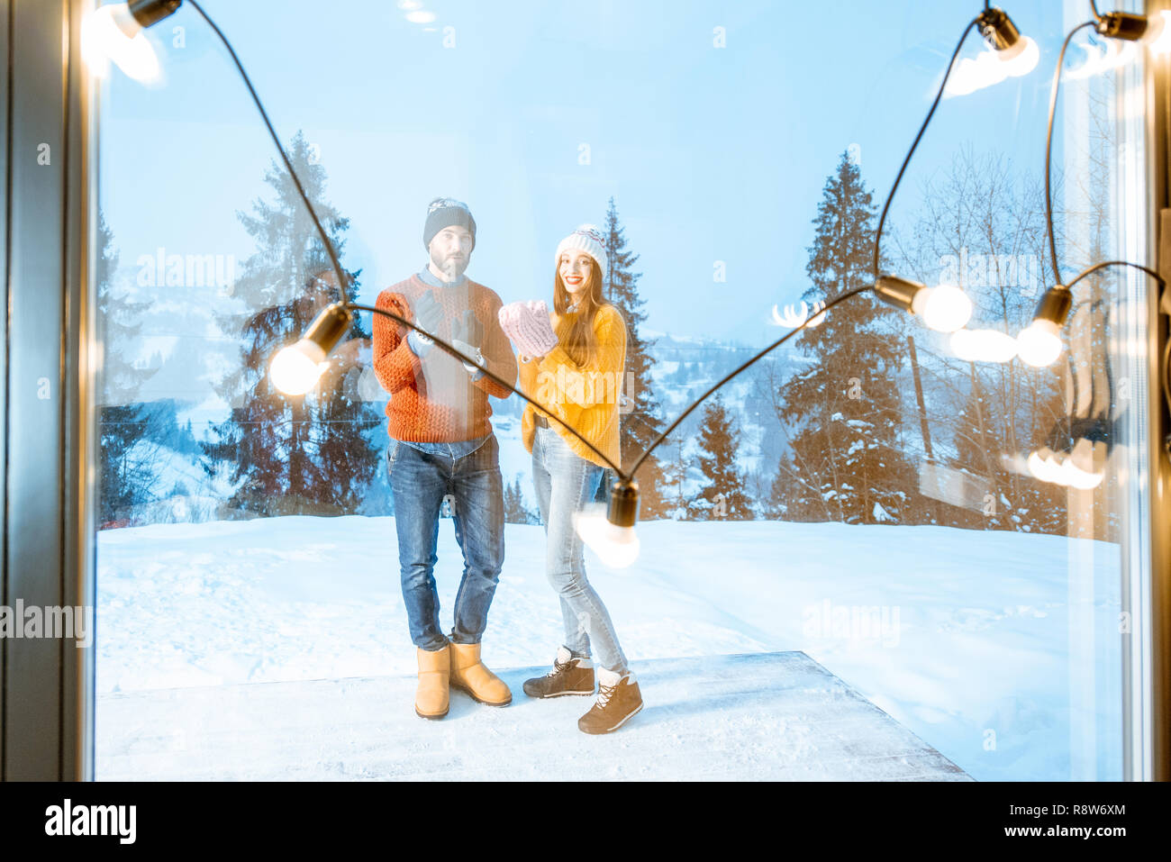 Coppia giovane in bright maglioni e cappelli in piedi sul terrazzo della casa in montagna durante il periodo invernale, vista attraverso la finestra con una ghirlanda Foto Stock