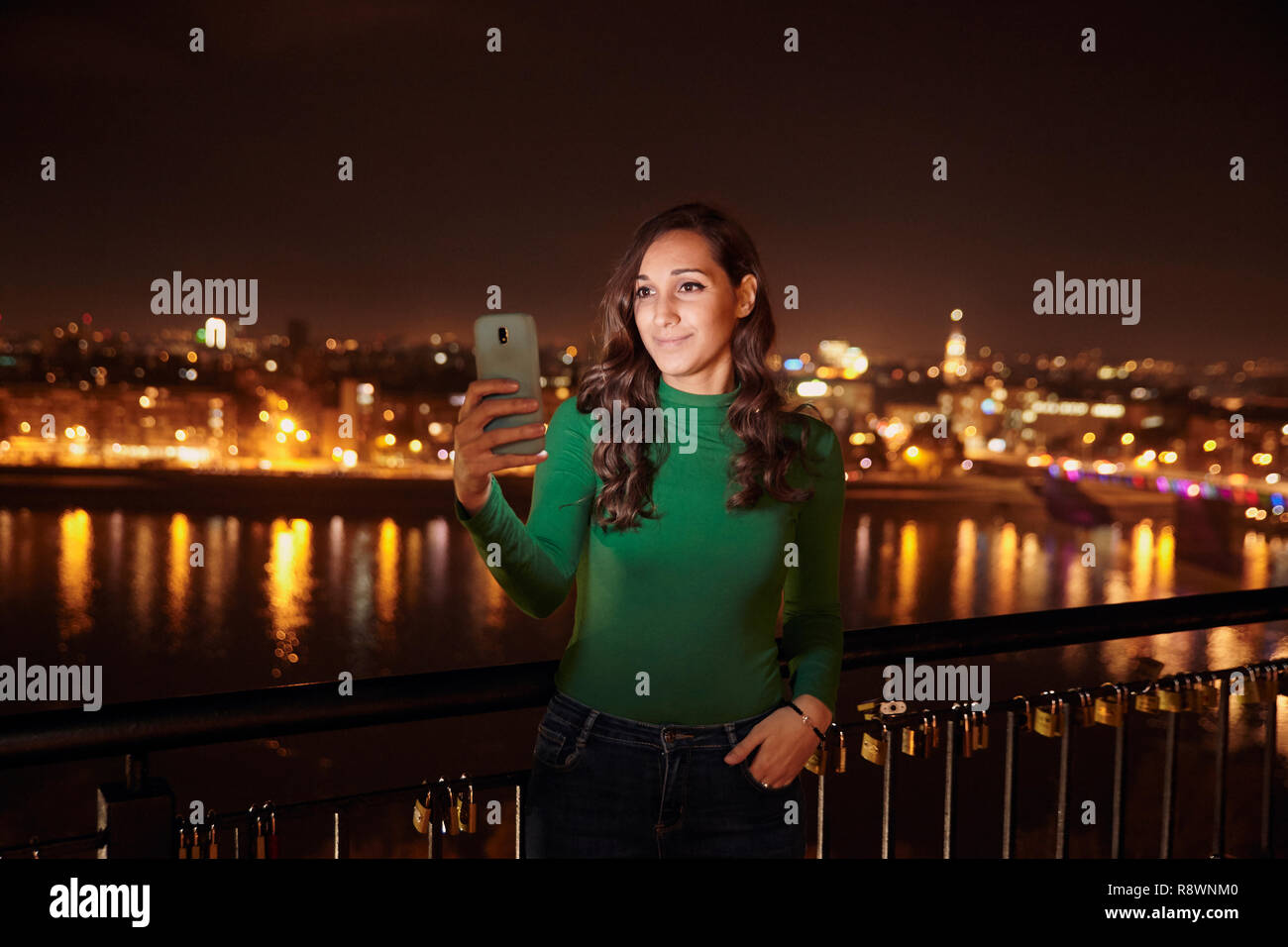 Una giovane donna bellissima, 20-25 anni, guardando a smart-phone. corpo superiore shot. Di notte, scuro, city scape dietro in background (sfocate, al di fuori del Foto Stock