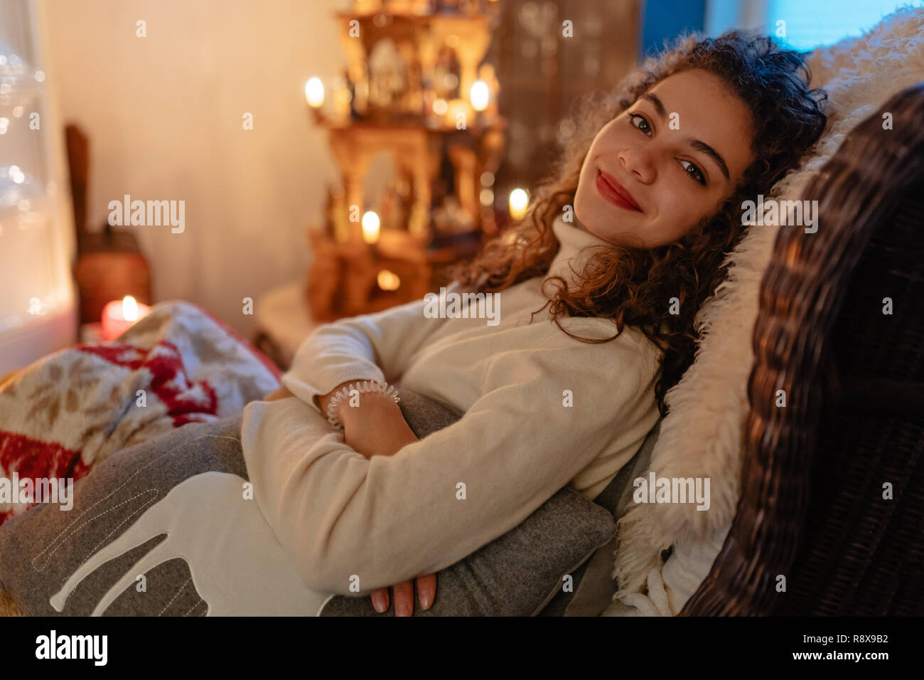 Bella sorridente giovane donna con capelli ricci è seduto rilassato su una grande sedia con decorazione di Natale in background Foto Stock