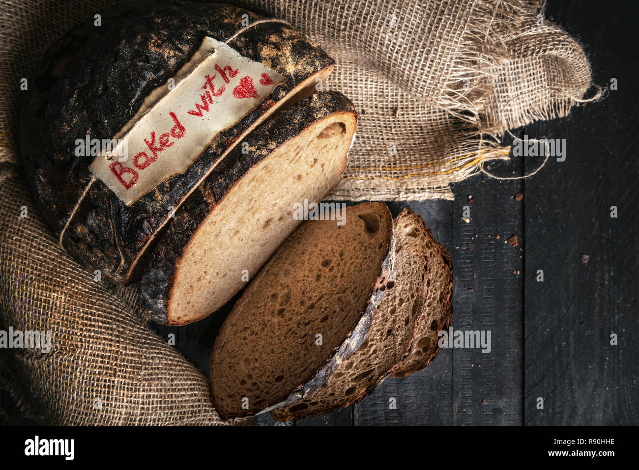 Pane appena sfornato sano pane marrone e cotto con cuore etiquette, sul sacco di iuta, su un rustico tavolo da cucina. Al di sopra di vista del tedesco a fette di pane di segale. Foto Stock