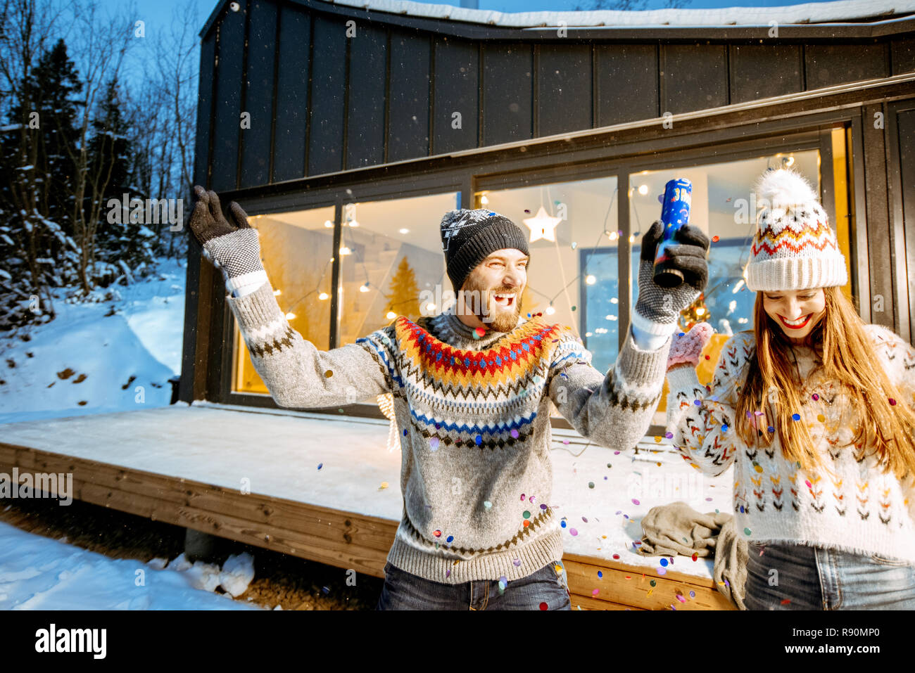 Giovane coppia felice vestito di maglioni per celebrare le vacanze invernali di fronte ad una bellissima casa decorata in montagna Foto Stock