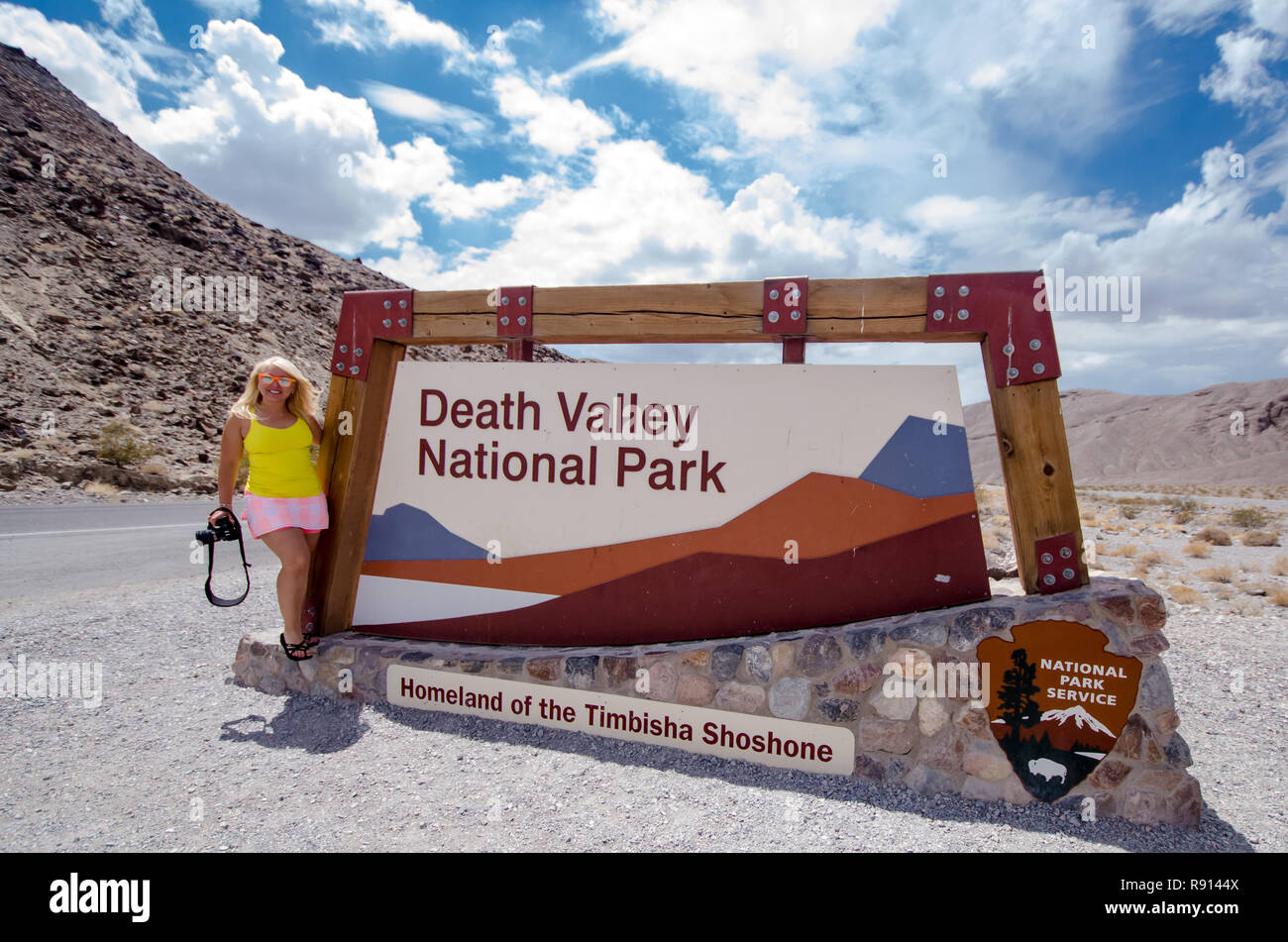 4 agosto 2018 - Death Valley, California: segno per il Parco Nazionale della Valle della Morte su un nuvoloso giorno d'estate.. Donna fotografo pone accanto a firmare Foto Stock