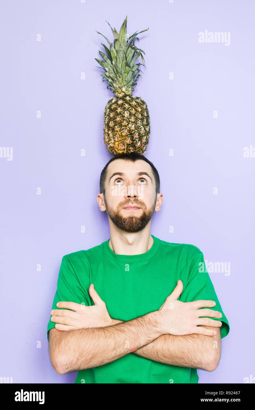 Giovane Azienda grande ananas sulla sua testa, braccia incrociate, guardando verso l'alto. Sfondo viola chiaro, copia spazio, orientamento verticale. Foto Stock