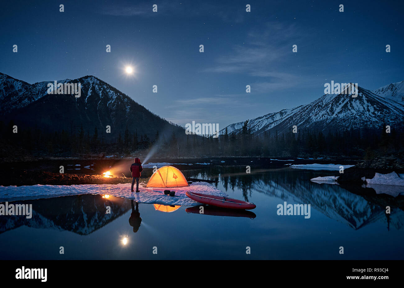 Vista della vita di camp. L'uomo avente un resto vicino al fuoco e arancio tenda accanto sul lago al chiaro di luna. Canoa vicino alla tenda del convegno. Foto Stock
