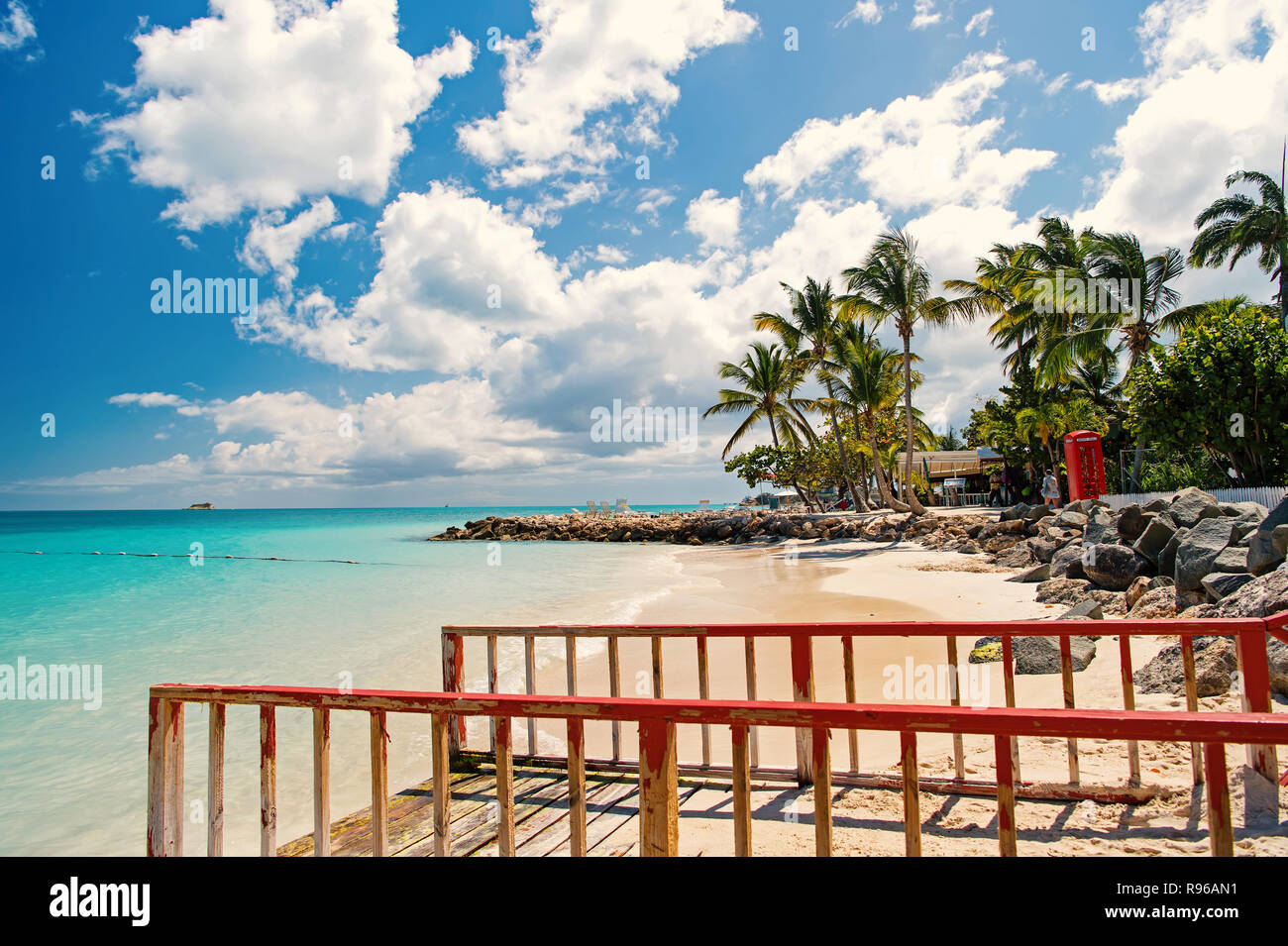 Pier con ringhiera in legno sulla spiaggia tropicale in st Johns, Antigua. Seascape con acqua turchese e verde delle palme, sabbia bianca sulla giornata di sole su nuvoloso cielo blu. La vacanza estiva concetto. Foto Stock