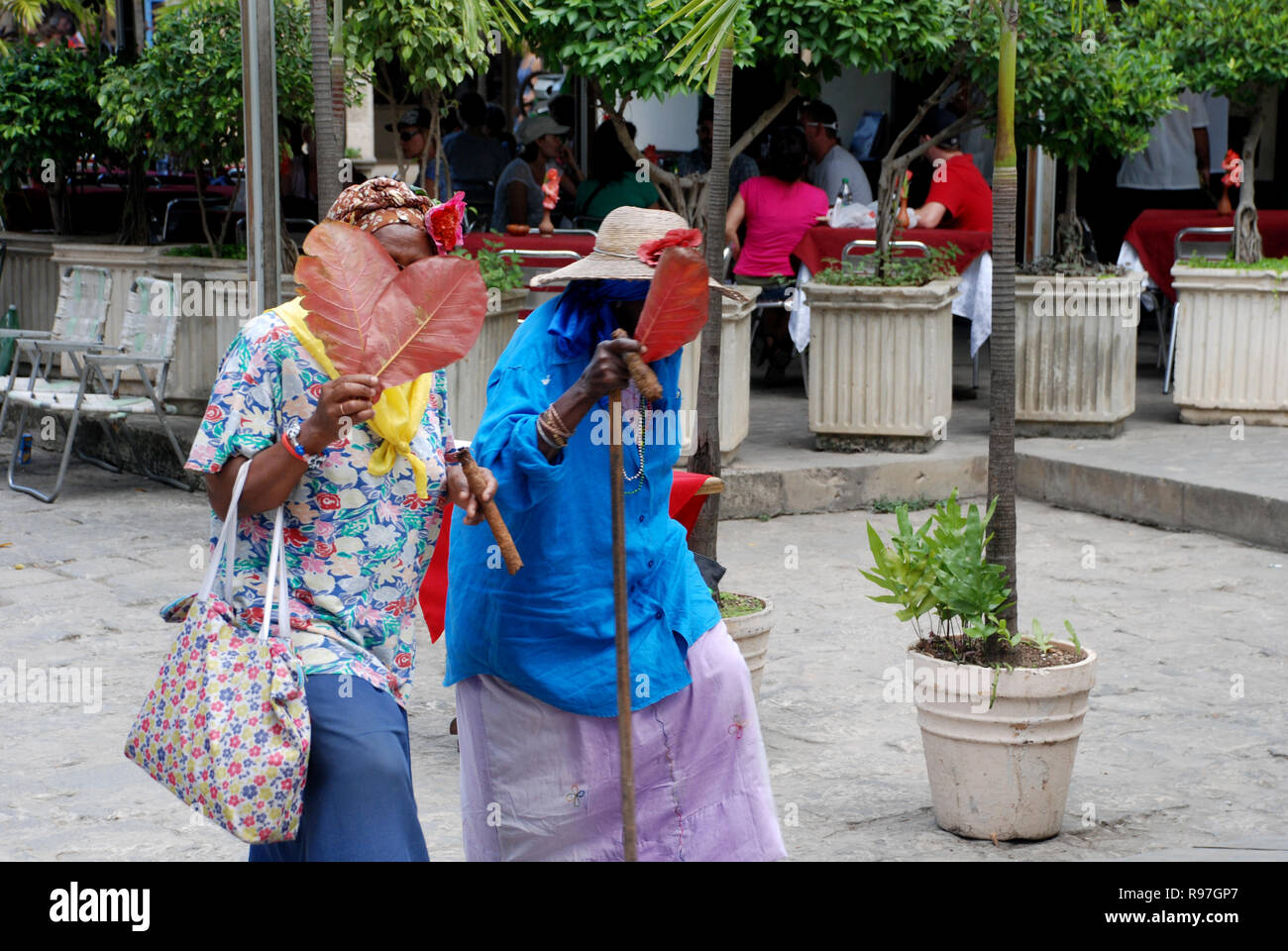 Due donne cubane in luminose vestiti a piedi su Havana con i sigari nelle loro mani, facce ricoperte con foglie di tabacco Foto Stock