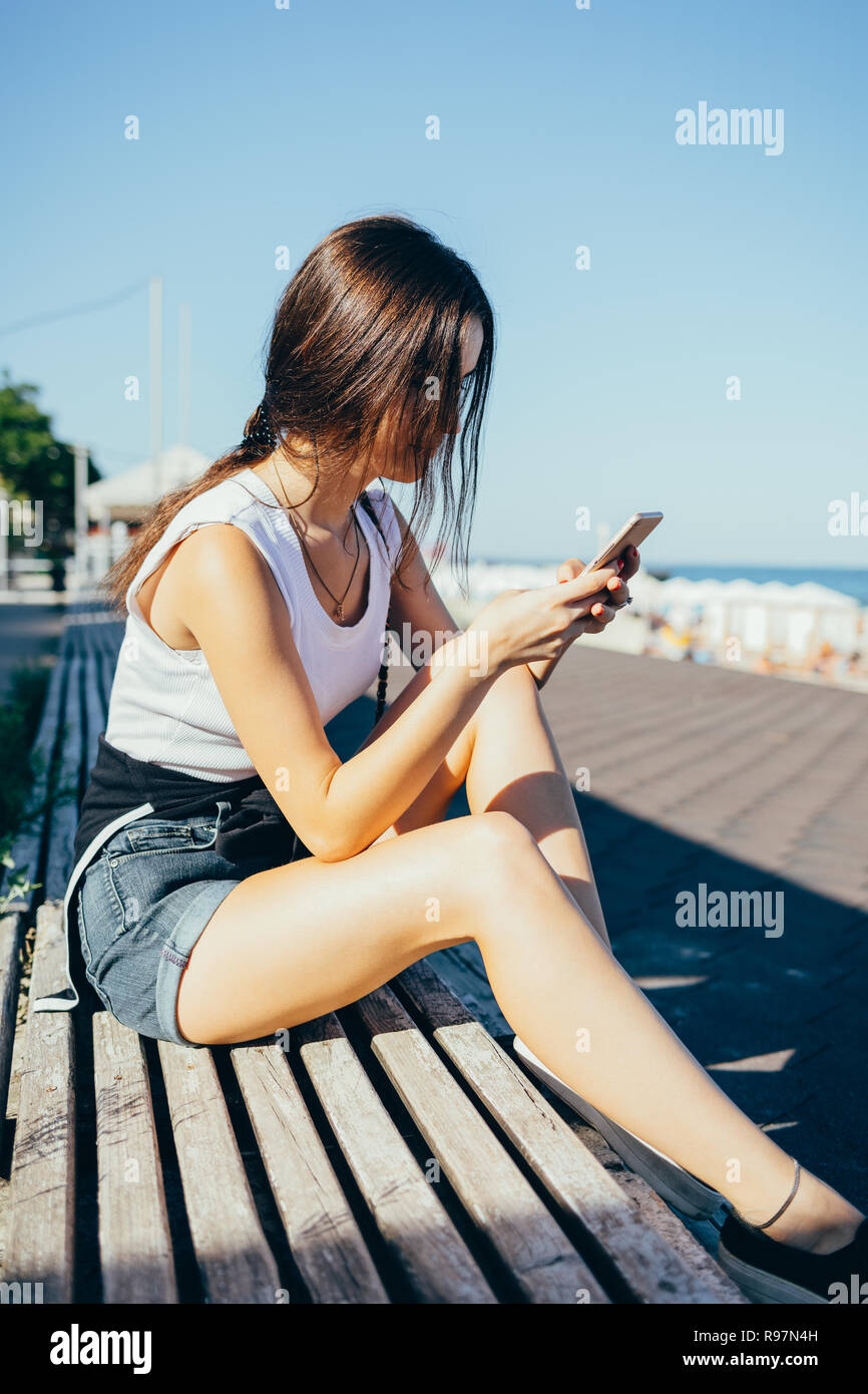 Giovane donna utilizzando smart phone all'esterno. Femmina bianca da indossare top e jeans corti seduto su estate mare guardando il dispositivo mobile. Foto Stock