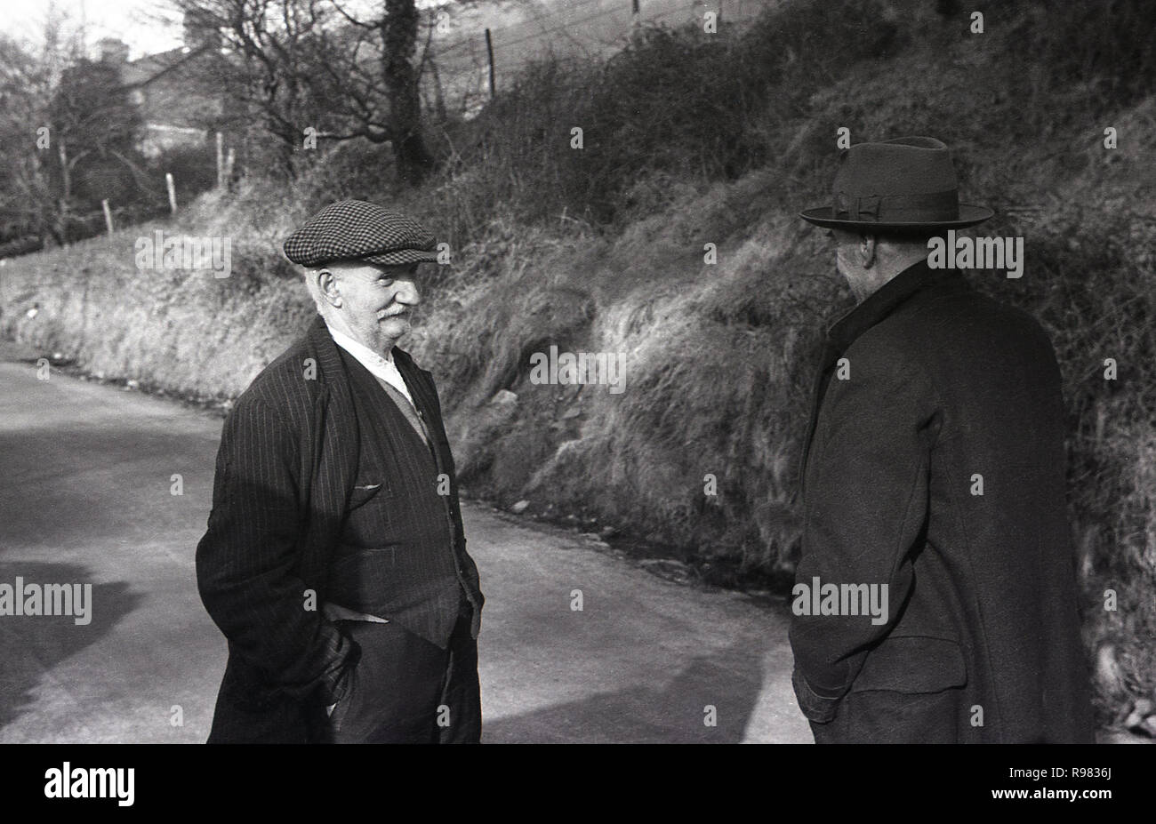 1940s, storico, due minatore gallese uno indossando un tappo piatto, altri un cappello, in piedi a una conversazione sulla strada di un paese al di fuori di Merthyr, Galles. Foto Stock